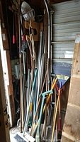 Overall view of the assortment of long-handled garden tools leaning against wall, including rakes, spades, and an electric trimmer.