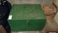 Closed green wooden tool chest with visible wood grain and metal latch, placed on a concrete floor by a wall.