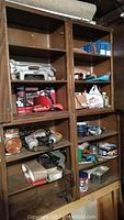 Wooden shelving unit with multiple power and hand tools, including boxed Craftsman cordless screwdriver kit, spray sprayer, drills, sanding tools, and cans.