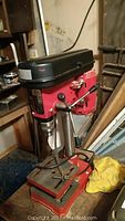 Side view of the red Brico bench drill showing the drill head, base, and metal work table with clamp.