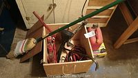 Wooden box filled with various automotive supplies including brooms, funnel, pry bar, jumper cables, and gas canister