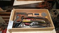 Top view of a wooden box containing various hand saws and accessories including a hacksaw, small saws with different colored handles, and metal blades.