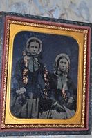 Close up of the photo depicting two girls in dresses and bonnets seated and standing, housed in a leather-bound case with a gold-tone tin frame and red velvet interior.
