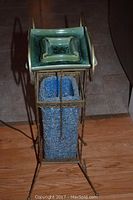 Full view of vintage ashtray light stand with twisted brass frame, blue textured internal plastic column, and green ceramic ashtray on top, standing on wooden floor.