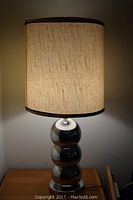 Lamp lit showing textured beige fabric shade with brown edge trim and chrome triple ball base.