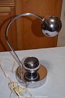 Front and full view of vintage chrome gooseneck eyeball desk lamp on a white surface, showing curved gooseneck and reflective spherical shade and base.