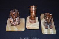 Three brass Egyptian figure sculptures mounted on rectangular cream marble bases, showing King Tutankhamun, Nefertiti bust, and Sphinx, against black background.
