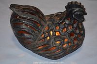 Black cast iron candle holder shaped like a chicken with hole cutouts and lit candle inside, showing glowing light.