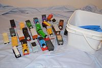 Multiple Thomas & Friends toy trains and freight cars arranged on fabric surface with white plastic bucket holding blue toy track pieces.