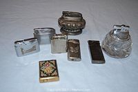 All seven vintage lighters arranged on white surface showing variety of styles and materials.