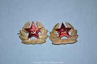 Two red star pins with gold-tone wheat designs and hammer and sickle symbols on blue background.