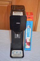 Front and side view of the SodaStream soda maker with the boxed CO2 carbonator standing beside it on a white table.