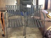 Photo showing two black cast iron patio chairs with vertical slats, armrests, and four legs with a slightly curved design.