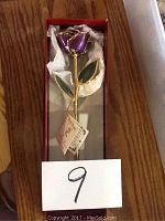 Photo of a real rose dipped in 24k gold with purple petals and green leaves, displayed inside a box with cushioning.
