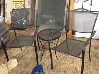 Photo showing two black metal patio chairs with mesh seats and backs and small round black metal table with mesh top
