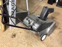 Gray MTD gas snow blower model 31A-142-033 with black handlebars and chute. White wheels on wooden floor.
