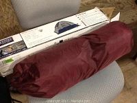 Photo showing the Wenzel Sport Dome Tent in a red carrying bag resting on a chair with its original box in the background.