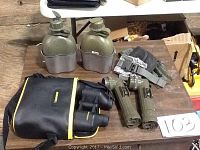 Photo showing the military gear on a wooden table: Bushnell binoculars in case, two olive green canteens with double cup holder, two olive green flashlights, and green military pouches.
