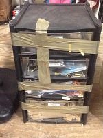 Black plastic four-drawer storage unit sealed with multiple tape strips holding drawers closed, drawers contain unknown items.