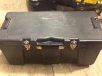 Photo of a closed rectangular black plastic tool chest placed on a floor, showing the top, front, and side portions including two metal latches and a central carrying handle.