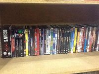 Full view of shelf containing approximately 30 assorted DVDs, mostly movie titles and TV series in standard DVD cases.