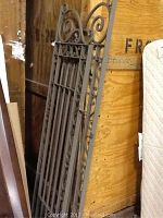 Metal gate leaning upright against wooden crate and mattress, showing detailed scroll design and vertical bars.
