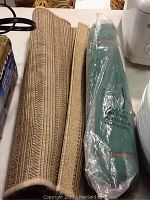 Two rolled mats on white surface: one beige woven outdoor mat and one green puzzle roll-up mat inside clear plastic.