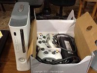 White Xbox 360 console standing upright next to an open box containing three white wired controllers and power adapter cable.