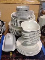 Stacked assortment of white ceramic dishes including plates, bowls, small casseroles, and a butter dish with lid.