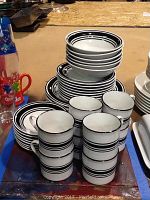 Photo showing stacks of white ceramic plates, bowls, and mugs with black stripe design on rims arranged on a surface.