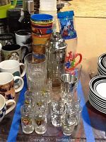 Photo showing clear glass shot glasses with cartoon character designs and metal rims, colorful kids plastic cups with Mickey Mouse and other characters, a stainless steel cocktail shaker, ceramic mugs with various designs, and partially visible plates with black trim.