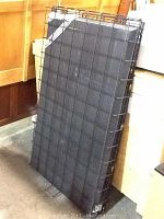 Photo of a black wire metal dog crate folded and leaning vertically against a wall, with a black base panel also visible.
