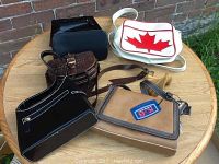 Five handbags arranged on a round wooden table outdoors next to a brick wall. Visible detail of material and design variety to show overall condition and style.