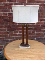 Full view of the table lamp against a brick wall on a wooden surface showing the white linen drum shade, wooden frame, and marble base.