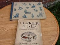 Photo showing two books, one with cream blue cover illustrations and one titled 'Currier & Ives: Printmakers to the American People' by Harry T. Peters, placed on a wooden table.