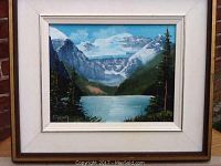 Full view of the framed oil painting showing the signature and date in the lower left corner, depicting Lake Louise with mountains, trees, and water.