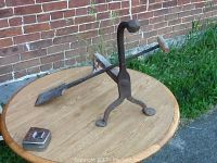 Photo of antique iron andiron and fireplace scraper placed on wooden table outdoors with brick wall background.
