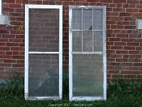 Two old storm windows side by side against a brick wall, one with a screen and one with glass panes, both with worn and chipped white wood frames.