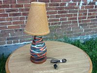 Overall view of the lamp showing the pottery base with colorful abstract patterns, wooden base, beige fabric shade, and electrical cord with plug.