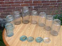 Eight glass preserve jars placed on a wooden surface against a brick wall, four glass lids in front.