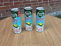 Three full vintage Quaker State Snowmobile Motor Oil cans placed upright on a wooden surface against a brick wall background, showing label fronts.