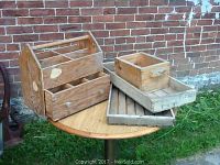 Wooden box with drawer and compartments and the two rectangular trays stacked with small box on top on a round wooden table outdoors against a brick wall.
