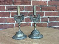Pair of brass-like candle holders standing on wooden surface in front of a brick wall showing full view of both pieces.
