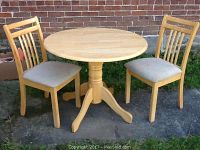 Full view of round maple kitchen table with two wooden chairs upholstered in beige fabric, showing the overall set arrangement and condition.