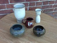Five handmade artisan pottery pieces on a wooden table in front of a brick wall. Includes vase, candle holder, pitcher, and two small bowls.