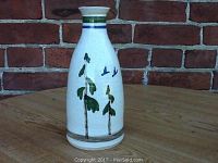 Front view of the white ceramic vase with green trees and blue birds painted on it, showing the narrow top and wide base against a wooden table and brick wall background.