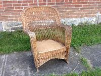 Front view of antique wicker rocking chair showing curved back, arms, and woven seat area on pavement against brick wall and grass.