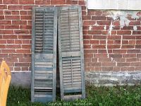 Two old wooden shutters with green paint leaning against a brick wall, dust and wear visible.