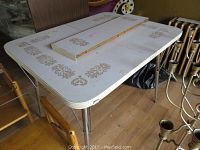 Full view of vintage folding table with decorative gold floral patterns, showing white laminated surface and metal legs, with removable leaves on top.