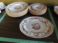 Photo showing bone china plates and platter, stacked and arranged on table with floral and pastoral motif in brown and multicolor flowers, gold scalloped edge.